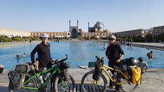 Zwei Frauen mit Hut und Sonnenbrille stehen mit ihrem bepackten Rädern vor dem Wasserbassin der Moschee in Isfahan, die im Hintergrund zu sehen ist.