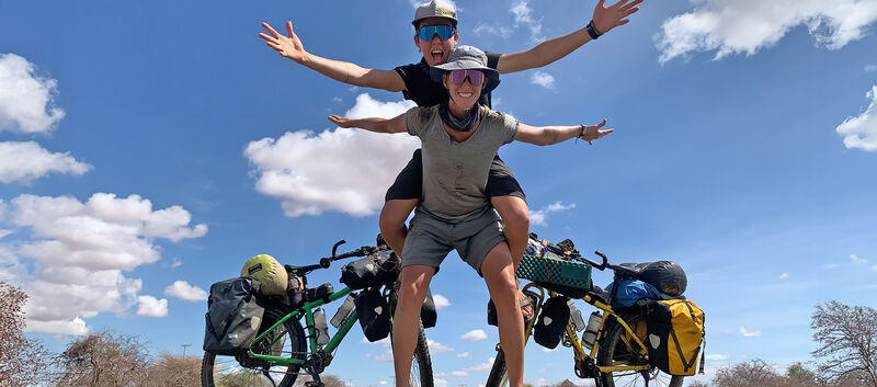 Die beiden Radabenteurerinnen Tanja Willers und Johanna Hochedlinger auf einer Straße in Sambia. Tanja sitzt auf dem Rücken von Johanna, beide stehen vor ihren Rädern.