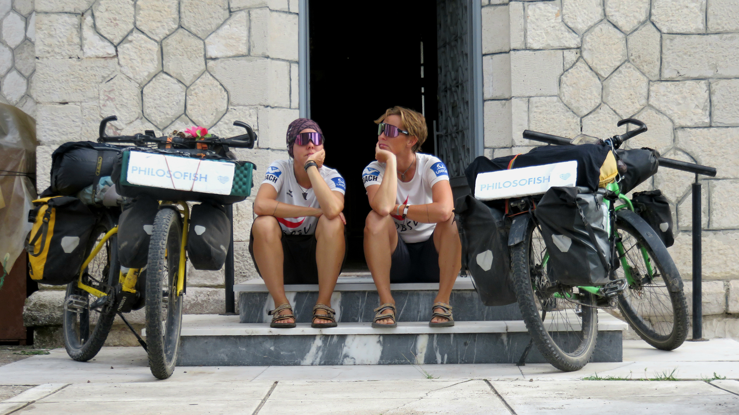 Zwei Frauen sitzen auf den Stufen vor einer offenen Tür, jeweils neben ihnen ihre Fahrräder mit Gepäck. 