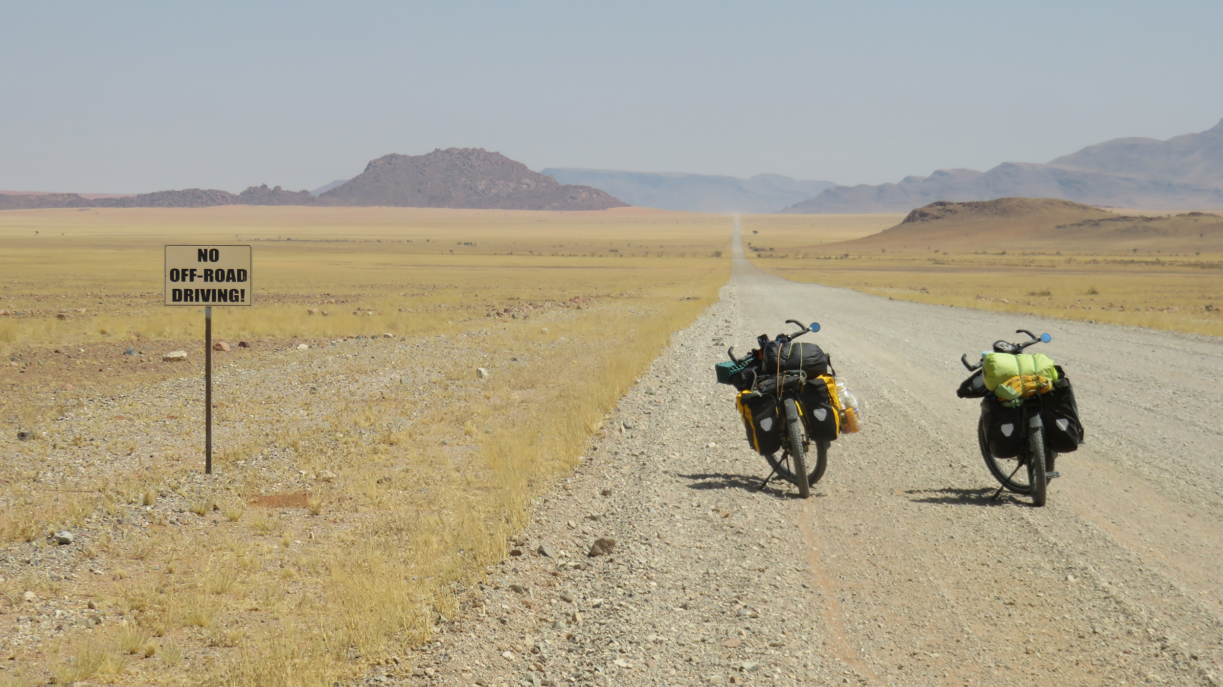Zwei mit Reisegepäck beladenen Fahrräder stehen auf einer Schotterstraße in Namibia vor der weiten Landschaft, im Hintergrund felsige Hügel.