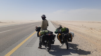 Eine Frau mit Hut und Mundschutz steht mit ihrem bepackten Fahrrad, daneben ein zweites an einer asphaltierten Straße. Daneben eine Ebene voll Wüstensand.