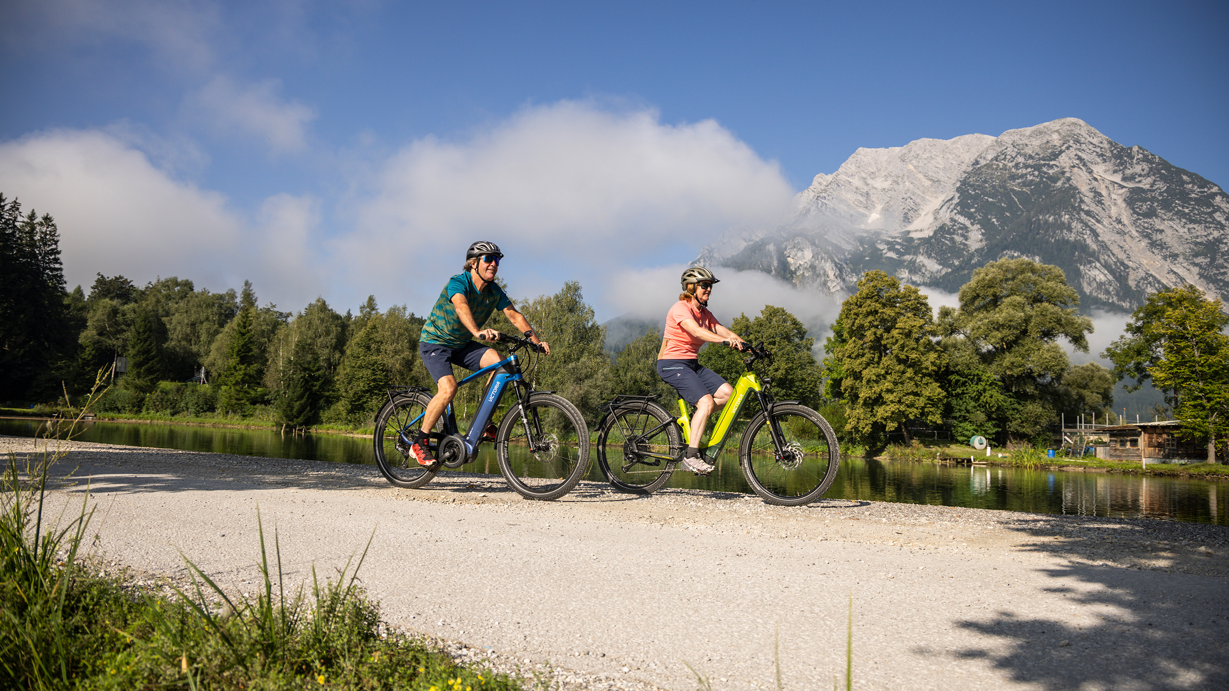 Zwei Radfahrer fahren auf einem Radweg an einem Fluss, im Hintergrund ein Berg