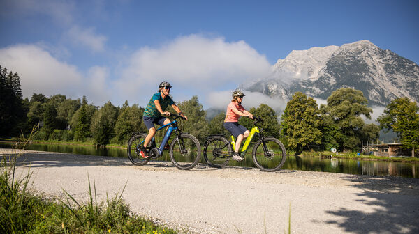 Zwei Radfahrer fahren auf einem Radweg an einem Fluss, im Hintergrund ein Berg