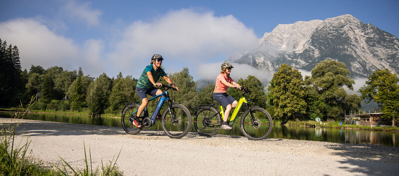 Zwei Radfahrer fahren auf einem Radweg an einem Fluss, im Hintergrund ein Berg