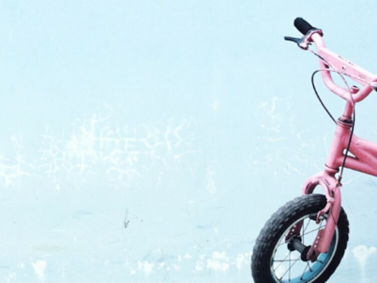 Header_Farrad_Kinder Auf dem Bild ist ein rosa Kinderfahrrad vor einem hellblauen Hintergrund zu sehen.