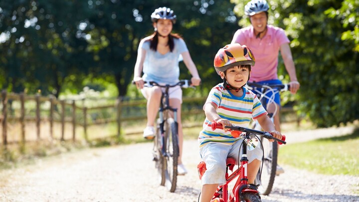 Fahrradausflug mit der Familie Auf dem Bild ist eine Familie bei beim Fahrradfahren zu sehen. Im Vordergrund fährt ein kleiner Junge, er trägt ein bunt gestreiftes T-Shirt und einen orangen Helm. Im Hintergrund, leicht verschwommen, sieht man die Mutter und den Vater auch jeweils auf einem Fahrrad.