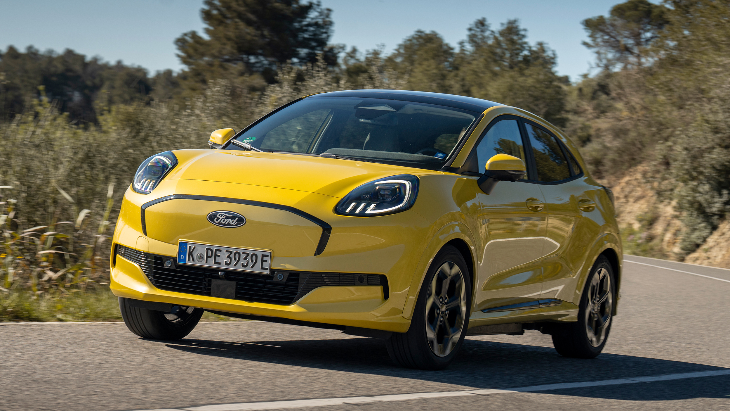 Ein gelber Ford Puma Gen-E fährt durch eine Kurve auf einer Landstraße.
