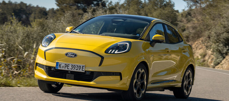 Ein gelber Ford Puma Gen-E fährt durch eine Kurve auf einer Landstraße.