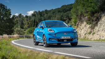 Ein blauer Ford Puma Gen-E fährt durch eine Kurve auf einer Landstraße.