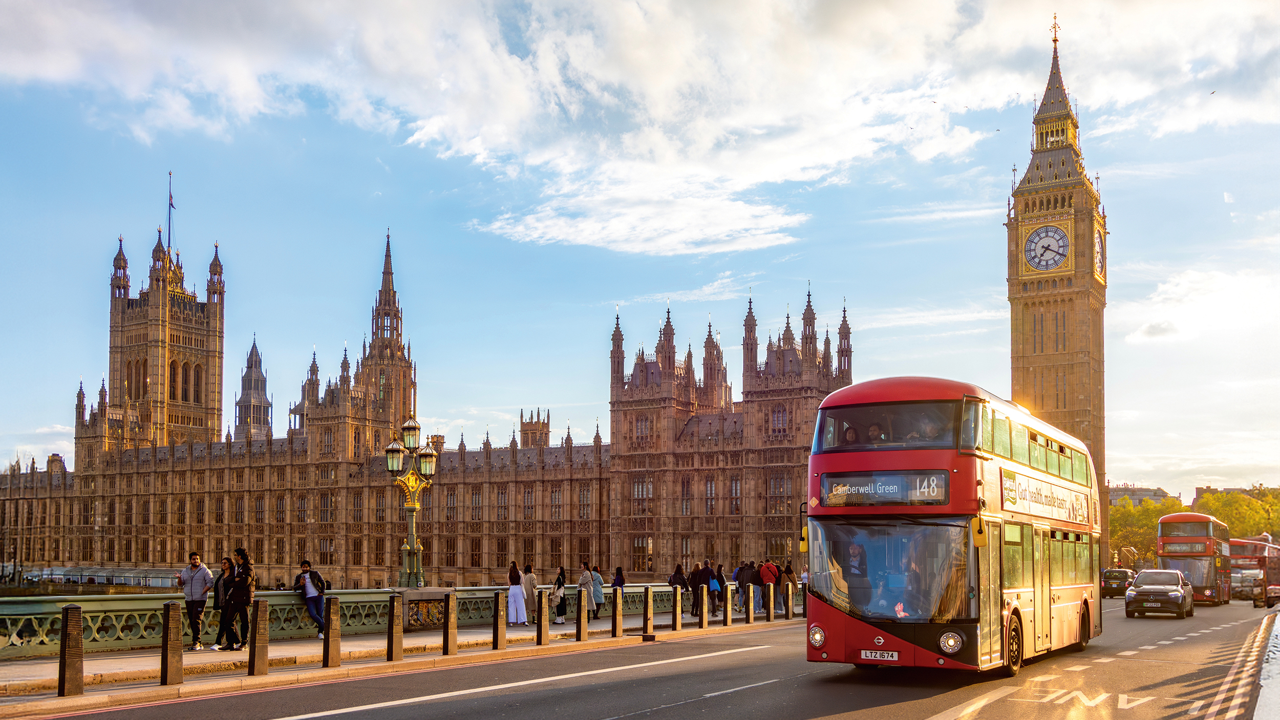Blick auf Big Ben von einer Brücke aus, es ist sonnig. Es fährt ein roter Bus vorbei.