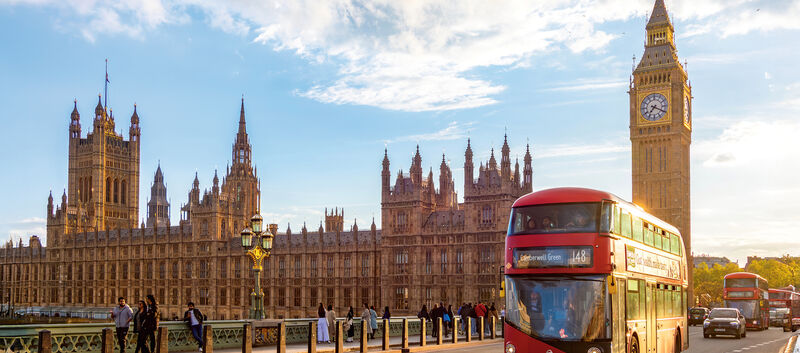 Blick auf Big Ben von einer Brücke aus, es ist sonnig. Es fährt ein roter Bus vorbei.