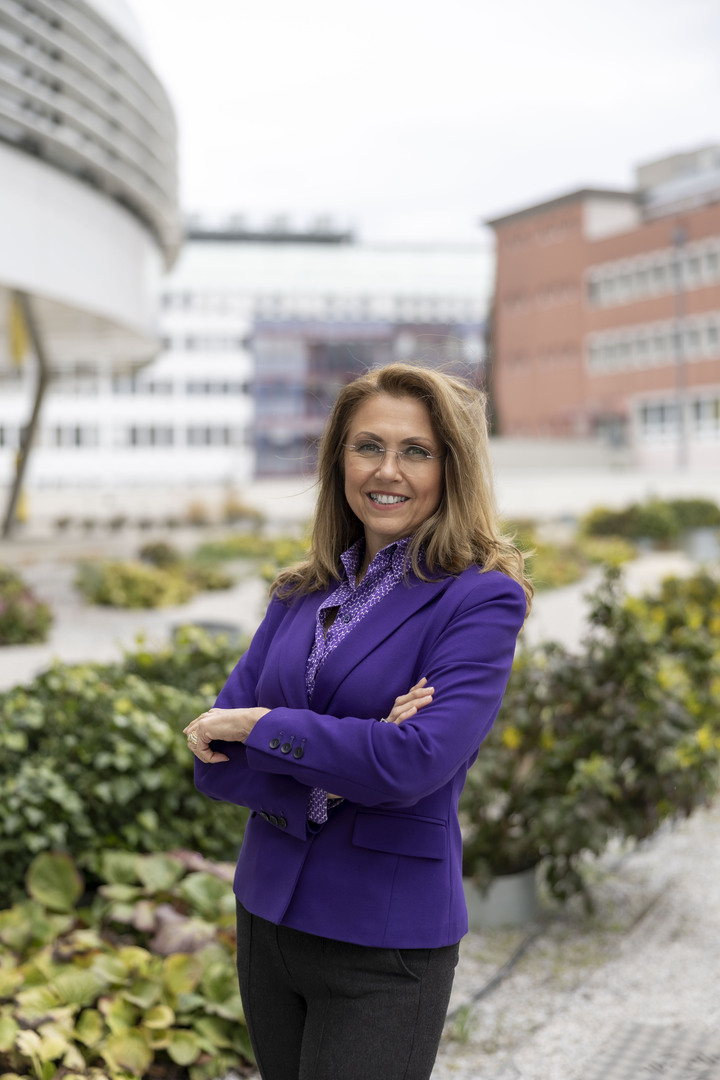 Auf dem Bild sieht man Marion Seidenberger mit verschränkten Armen. Sie trägt einen violetten Blazer und eine schwarze Hose. Sie schaut in die Kamera und lächelt. Im Hintergrund sieht man grüne Pflanzen.