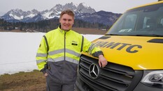 Auf dem Bild sieht man einen jungen Pannenfahrer des ÖAMTC. Er steht neben seinem gelben Pannenfahrzeug und schaut in die Kamera. Er hat eine graue Hose und eine gelbe Jacke an. Im Hintergrund sieht man eine Wiese, die leicht schneebedeckt ist. In einiger Entfernung kann man das Tiroler Bergpanorama erahnen.