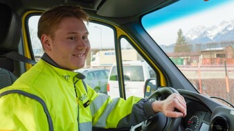 Auf dem Bild sieht man einen jungen Pannenfahrer des ÖAMTC. Er sitzt am Lenkrad seines Pannenfahrzeugs und schaut auf die Straße. Er hat eine gelbe Jacke an. Im Hintergrund sieht man einige geparkte Autos. In einiger Entfernung kann man das Tiroler Bergpanorama erahnen.