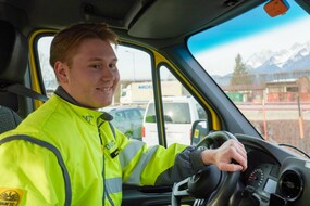 Auf dem Bild sieht man einen jungen Pannenfahrer des ÖAMTC. Er sitzt am Lenkrad seines Pannenfahrzeugs und schaut auf die Straße. Er hat eine gelbe Jacke an. Im Hintergrund sieht man einige geparkte Autos. In einiger Entfernung kann man das Tiroler Bergpanorama erahnen.