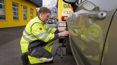 Auf dem Bild sieht man einen jungen Pannenfahrer des ÖAMTC. Er hockerlt neben einem dunkelgrauen Pkw undhantiiert am Vorderreifen des Fahrzeugs. Er hat eine graue Hose und eine gelbe Jacke an. Im Hintergrund sieht man einen gelben ÖAMTC Stützpunkt.