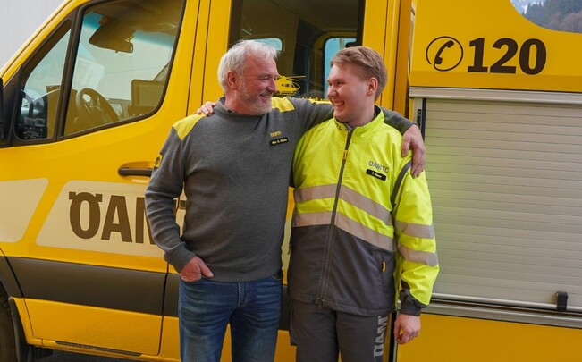 Auf dem Bild sieht man einen jungen sowie einen älteren Pannenfahrer des ÖAMTC. Beide stehen vor einem gelben Pannenfahrzeug. Sie umarmen sich freundschaftlich und schauen einander freundlich an. Der ältere trägt eine blaue Jean und einen grauen Pullover mit ÖAMTC Schriftzug. Der jüngere hat eine graue Hose und eine gelbe Jacke ebenfalls mit ÖAMTC Logo an.