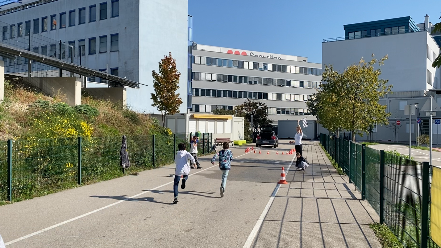 Auf dem Bild sieht man zwei Kinder, die schnell eine Fahrbahn entlanglaufen. Im Hintergrund steht eine Frau mit erhobenem Arm. Die Fahrbahn ist von einem grünen Zaun begrenzt. Am Ende der Fahrbahn steht eine Reihe orangener Verkehrskegel. Am Rand der Fahrbahn steht ein orange-weiß gestreifter Verkehrskegel.