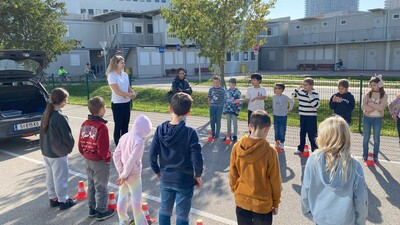 Auf dem Bild sieht man zwei Reihen Kinder, die eine Asphaltierte Fahrbahn flankieren. Die Kinder sind bunt gekleidet. Auf der Fahrbahn steht eine Frau mit schwarzer Hose und weißem T-Shirt. Sie erklärt den Kindern etwas. Vor jedem Kind steht ein kleiner orange-weiß gestreifter Verkehrskegel.