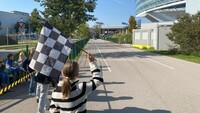 Auf dem Bild sieht man ein Mädchen von hinten. Das Mädchen hat einen Zopf und trägt einen schwarz-weiß gestreiften Pulli. Über ihrem Kopf schwingt sie eine schwar-weiß karrierte Fahne. Sie steht am Rand einer asphaltierten Fahrbahn. Im Hintergrund kann man ein schwarzes Auto erkennen. Seitlich der Fahrbahn sitzen noch andere Kinder.