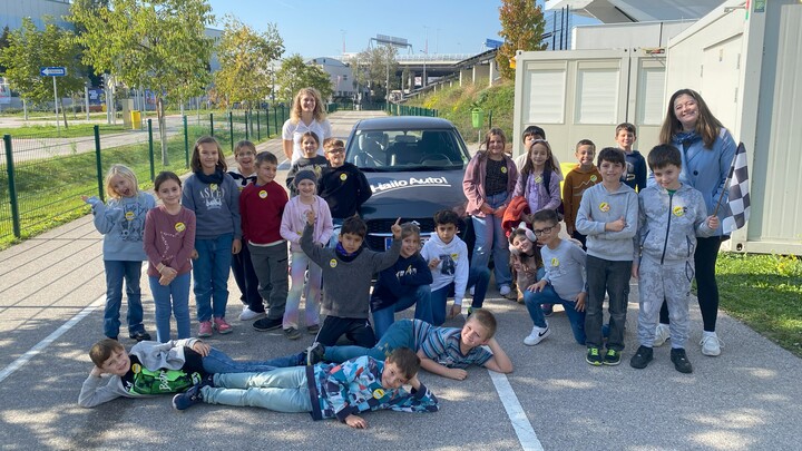 Auf dem Bild sind man eine Gruppe Volksschulkinder. Sie stehen, sitzen und liegen rund um ein schwarzes Auto. Auf der Motorhaube des Autos steht in weißer Schrift Hallo Auto. Die Kinder schauen in die Kamera und lachen. Auch zwei erwachsene Begleiterinnen sind zu sehen. Im Hintergrund kannn man eine grüne Böschung, einen grünen Zaun, sowie eine Brücke aus Beton erkennen. Das Auto steht auf eines aspahltierten Straße.Vor hellen Wohncontainern.