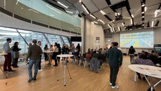 Auf dem Bild sieht man zahlreiche Personen sitzend und stehend in einem Saal.Im Vordergrund stehen einige Stehtische. Auf der linken Seite hat der Saal eine Fensterfront. Im Hintergrund kann man eine Leinwand mit einem Präsentationschart zum 15. Innovationsfrühstück erkennen. Der Fußboden besteht aus braunem Parkett.