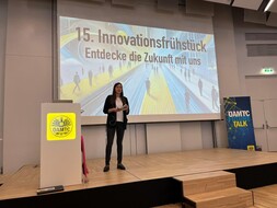 Auf dem Bild sieht man eine junge Frau. Sie steht auf einer Bühne. Sie trägt ein weißes T-Shirt, schwarze Hode und einen schwarzen Blazer. Links neben ihr steht ein Rednerpult mit dem gelben ÖAMTC Logo. Recht im Hintergrund ein Rollup mit der Aufschrift "Öamtc Future Talk". Hinter ihr auf der großen Präsentaionsleinwand ist der Schriftzug "15. Innovationsfrühstück - Entdecke die Zukunft mit uns" zu lesen.