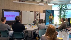 Auf dem Bild sieht man einen modern eingerichteten Schulungsraum. Um Tische die mit Wasserkrügen und Wassergläsern gedeckt sind, sitzen insgesamt sechs Personen, von denen man nur die Rücken sieht. Zwei Frauen stehen vor ihnen und moderieren eine Veranstaltung. Auf dem großen Monitor auf der linken Seite des Bildes erkennt man den Titel der Veranstaltung: "Open Space Days, Trendscouting."