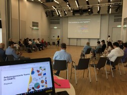 Auf dem Bild sieht man mehrere Personen in einem großen, fensterlosen Saal. An der Rückwand des Saal ist eine Leinwand angebracht, auf der gerade eine Präsentation zu sehen ist. Im Vordergrund sieht man den Bildschirm eines Laptops mit der aktuellen Folie. Auf dieser kann man den Titel der Präsentation lesen: "Selbstorganisierte Teams im ÖAMTC."