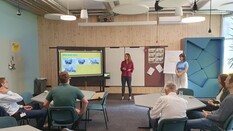 Auf dem Bild sieht man eine Frau in einem modern eingerichteten Schulungsraum stehen. Sie steht vor einem großen Bildschirm, auf dem Teile einer Präsentation zu erkennen sind. Die Frau hat ein weinrotes Oberteil und eine schwarze Hode an. Insgesamt hören ihr fünf sitzende und eine stehende Person zu.