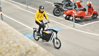 Von schräg oben fotografiertes, fahrendes, einspuriges Lastenfahrrad mit gefüllter Gepäcktasche, die vor dem Lenker montiert ist