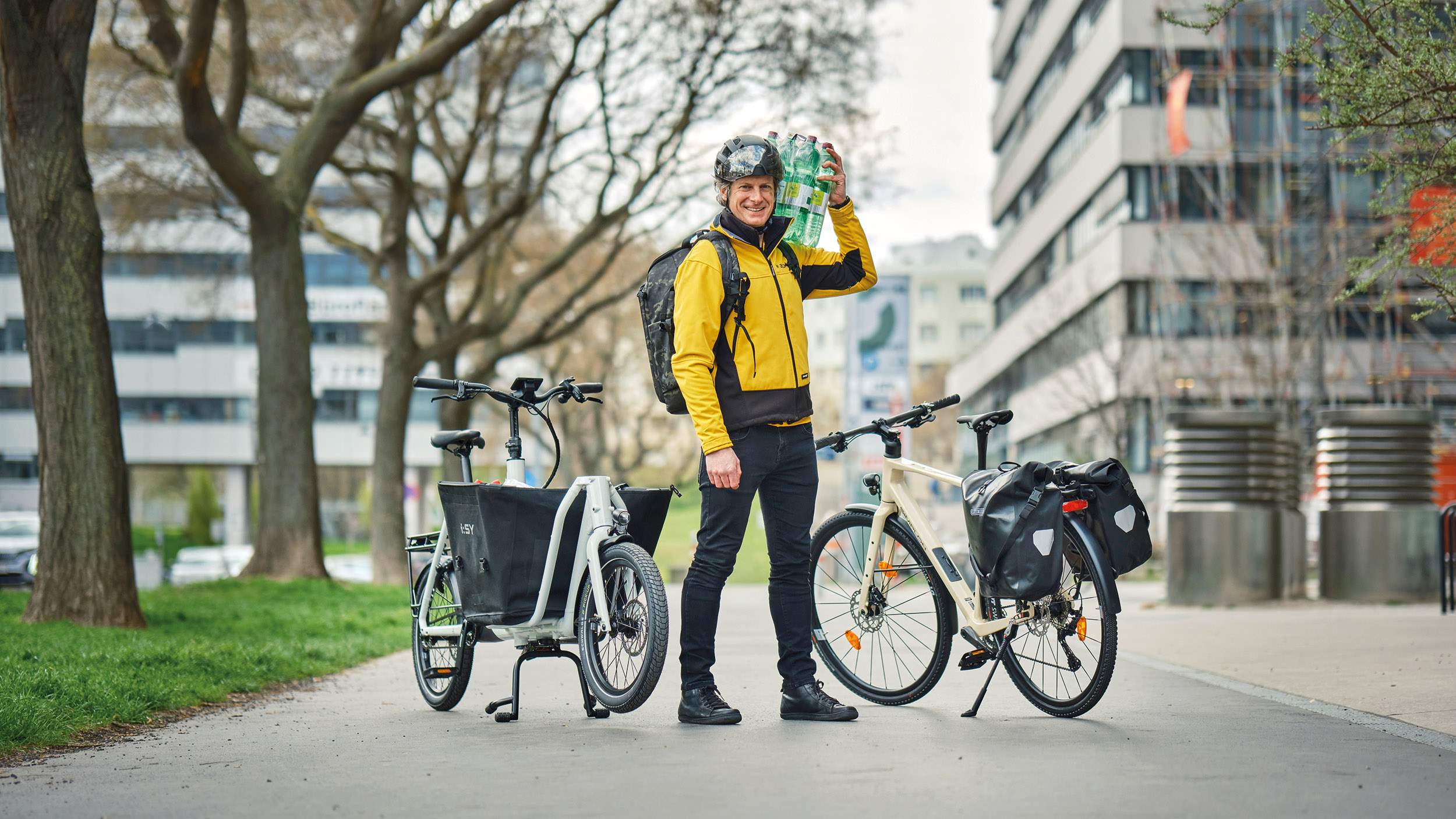 Redakteur mit gelber Jacke und geschulterten Wasserflaschen steht zwischen einem Lastrad und einem Fahrrad mit Gepäcktaschen.