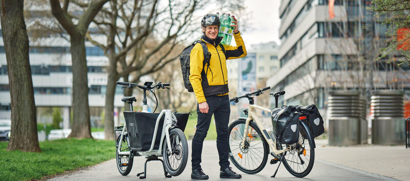 Redakteur mit gelber Jacke und geschulterten Wasserflaschen steht zwischen einem Lastrad und einem Fahrrad mit Gepäcktaschen.