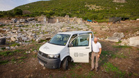 Archäologe Simon Hye steht vor dem weißen VW-Bus, mit dem er nach Ephesos gefahren ist. Im Hintergrund sind Ruinen zu sehen.