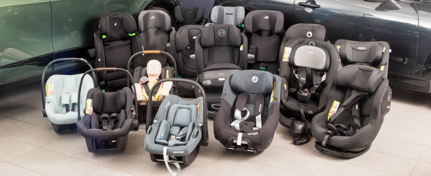 16 Kindersitze in unterschiedlichen Größen und Farben stehen vor zwei Autos am Boden einer Testhalle.