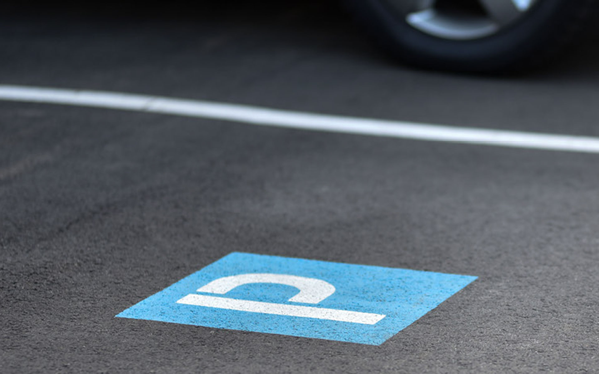Kennzeichnung eines Parkplatzes mit dem Symbol P am Boden (weiß auf blauem Hintergrund) und weißer Randlinie; hinter der weißen Linie sieht man ein geparktes Auto.