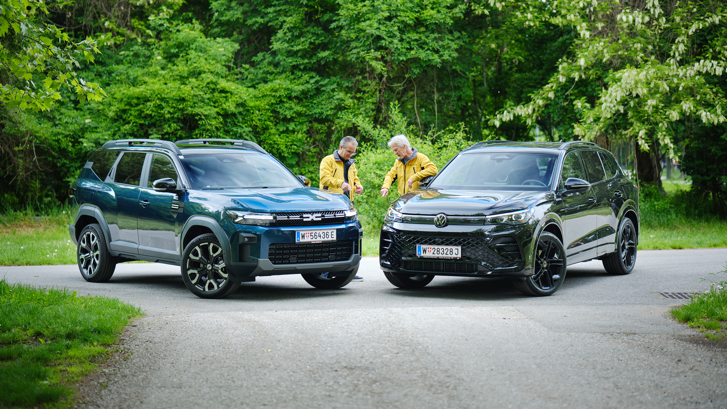 Dacia Bigster und VW Tiguan von schräg vorne, dazwischen stehen zwei Testredakteure