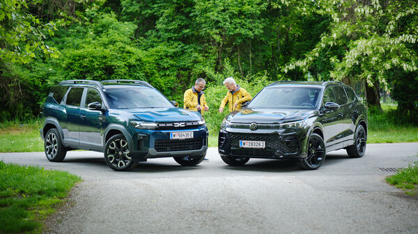 Dacia Bigster und VW Tiguan von schräg vorne, dazwischen stehen zwei Testredakteure