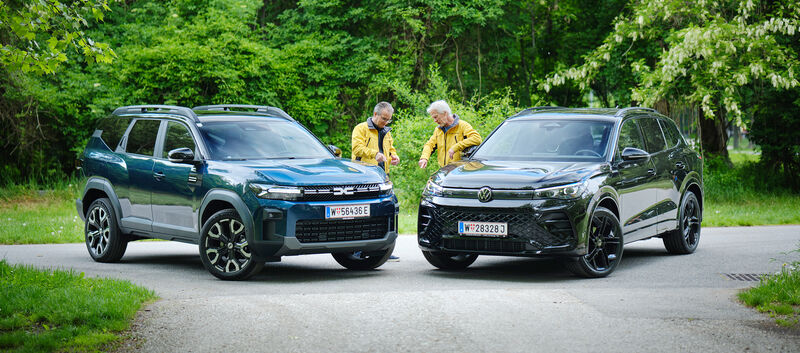 Dacia Bigster und VW Tiguan von schräg vorne, dazwischen stehen zwei Testredakteure