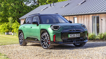 Ein grüner Mini JCW Aceman steht auf einem Schotterplatz vor einem Holzhaus.