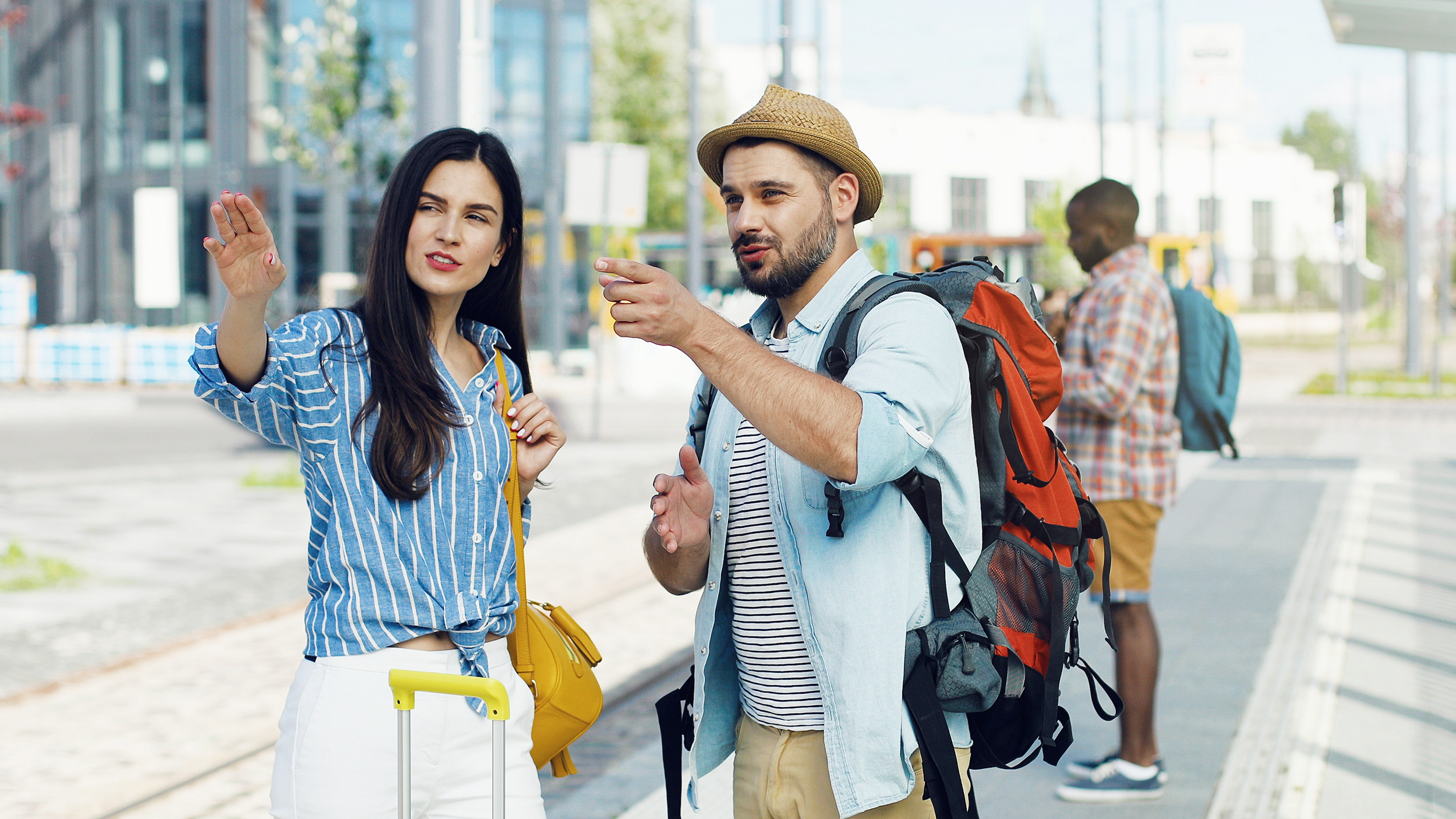 Caucasian young beautiful woman tourist standing at bus stop with suitcase. Handsome male traveller with backpack coming to ask the route. Man asking for right way Girl explaining how to get somewhere