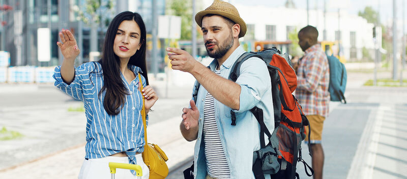 Caucasian young beautiful woman tourist standing at bus stop with suitcase. Handsome male traveller with backpack coming to ask the route. Man asking for right way Girl explaining how to get somewhere