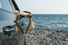Auf den Bild sieht man die die Seitenansicht der Beifahrerseite eines Autos. Das Auto steht auf einem Kiesstrand am Meer. Das Seitenfenster ist offen. Die Hand einer Frau, die einen Strohhut hält, hängt heraus.