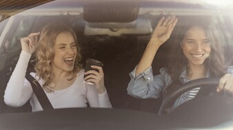 Auf dem Bild sieht man zwei junge Frauen durch die Windschutzscheibe eines Autos. Die rechte ist dunkelhaarig, trägt eine blaue Bluse und lenkt das Fahrzeug. Die linke hat helle Haare und ein weißes, langarmiges Shirt. Beide scheinen zu singen.