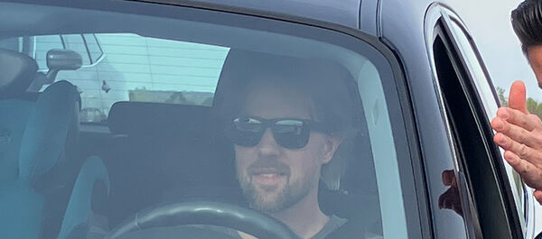 Auf dem Bild sieht man einen Mann mit Bart und schwarzer Sonnenbrille durch die Windschutzscheibe eines Autos. Neben dem Auto steht ein weiterer Mann, der ihm etwas erklärt.