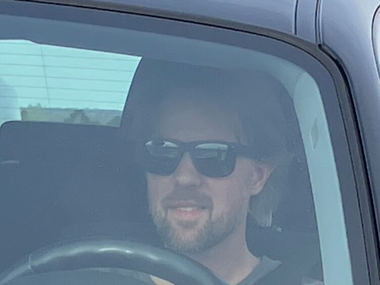 Auf dem Bild sieht maneinen Mann mit Bart und schwarzer Sonnenbrille durch die Windschutzscheibe eines Autos. Neben dem Auto steht ein weiterer Mann, der ihm etwas erklärt.