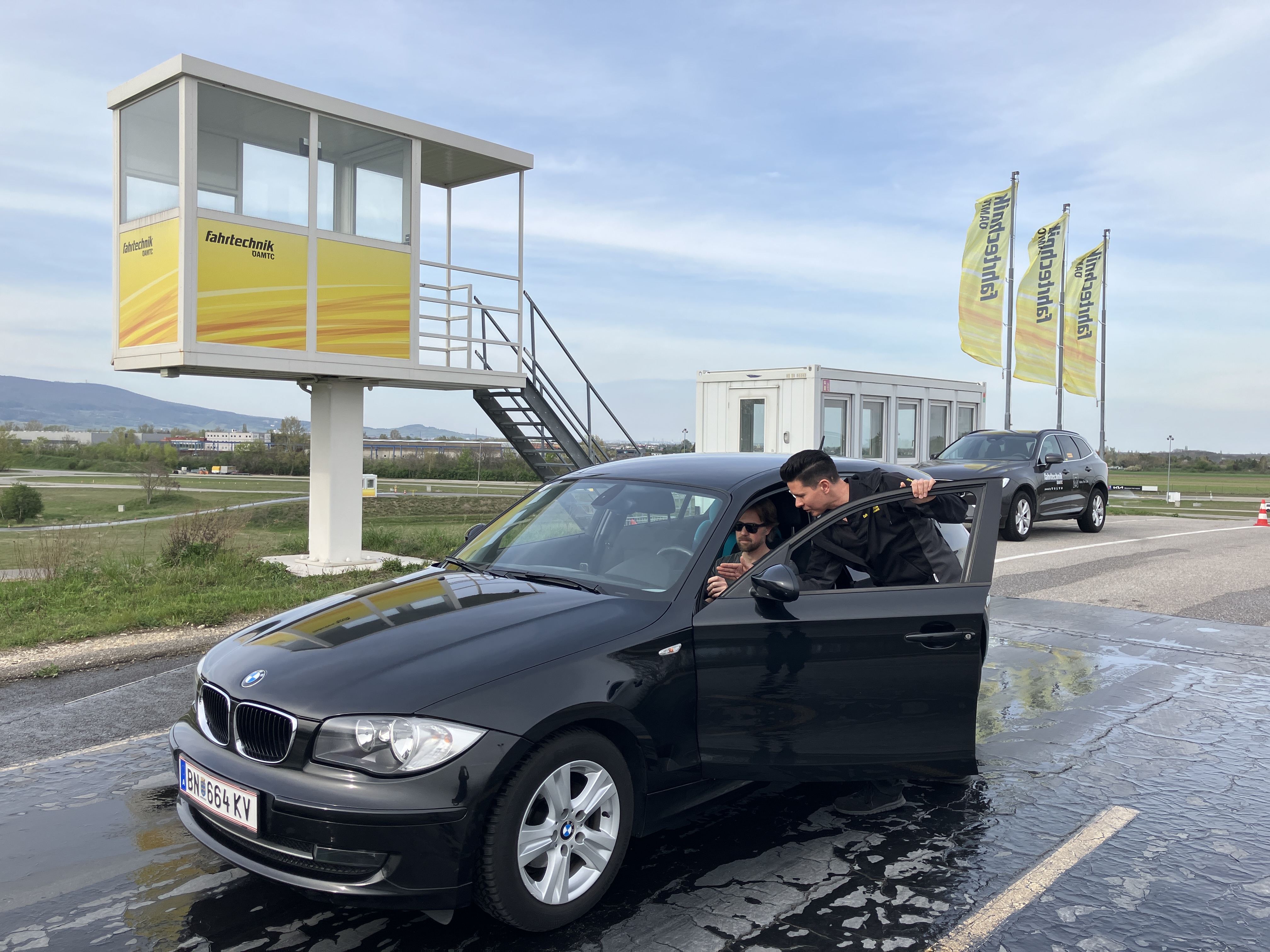 Auf dem Bild sieht man ein schwarzes Auto. Es steht auf einer feuchten Fahrbahn. Im Auto sitz eine Person. Die Fahrertür ist geöffnet. Daneben steht ein Mann der mit dem Lenker des Fahrzeugs spricht. Im Hintergrund sieht man eine etwas erhöhte weiße Kabine auf der Fahrtechnik steht. Rechts im Hintergrund wehen drei gelbe Fahnen.