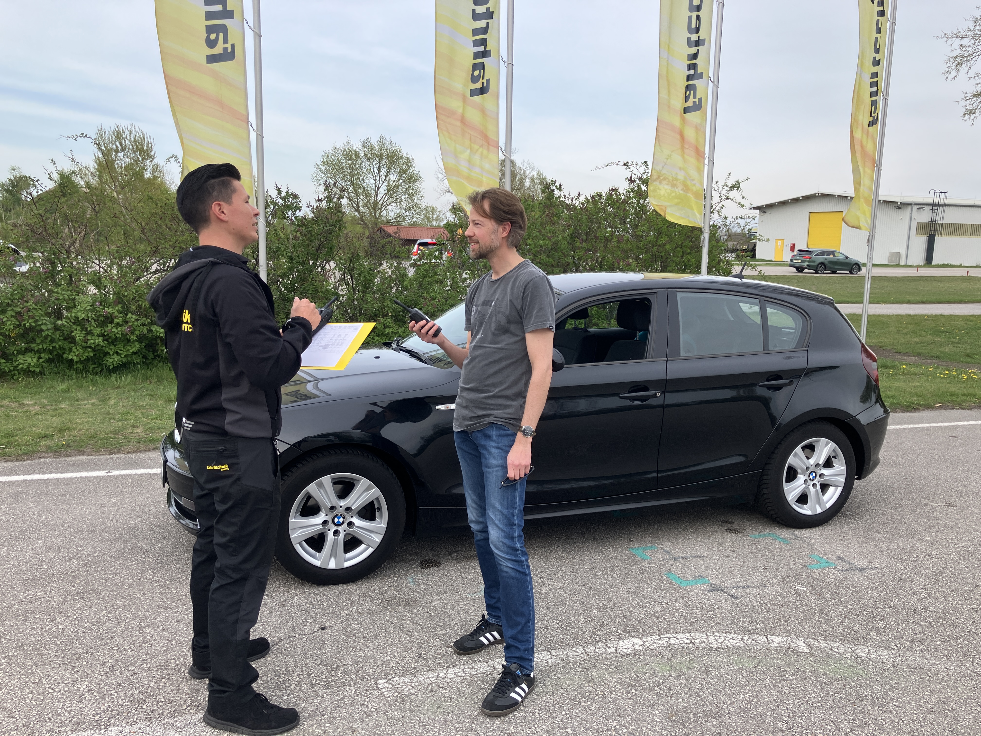 Auf dem Bild sieht man zwei Männer vor schwarzen einem Auto. Beide sind dunkel gekleidet. Der linke ganz in schwarz, der rechte in blauer Jeans und grauem Leiberl. Die beiden stehen sich gegenüber und reden miteinander. Im Hintergrund wehen vier gelbe Fahnen auf denen Fahrtechnik steht.