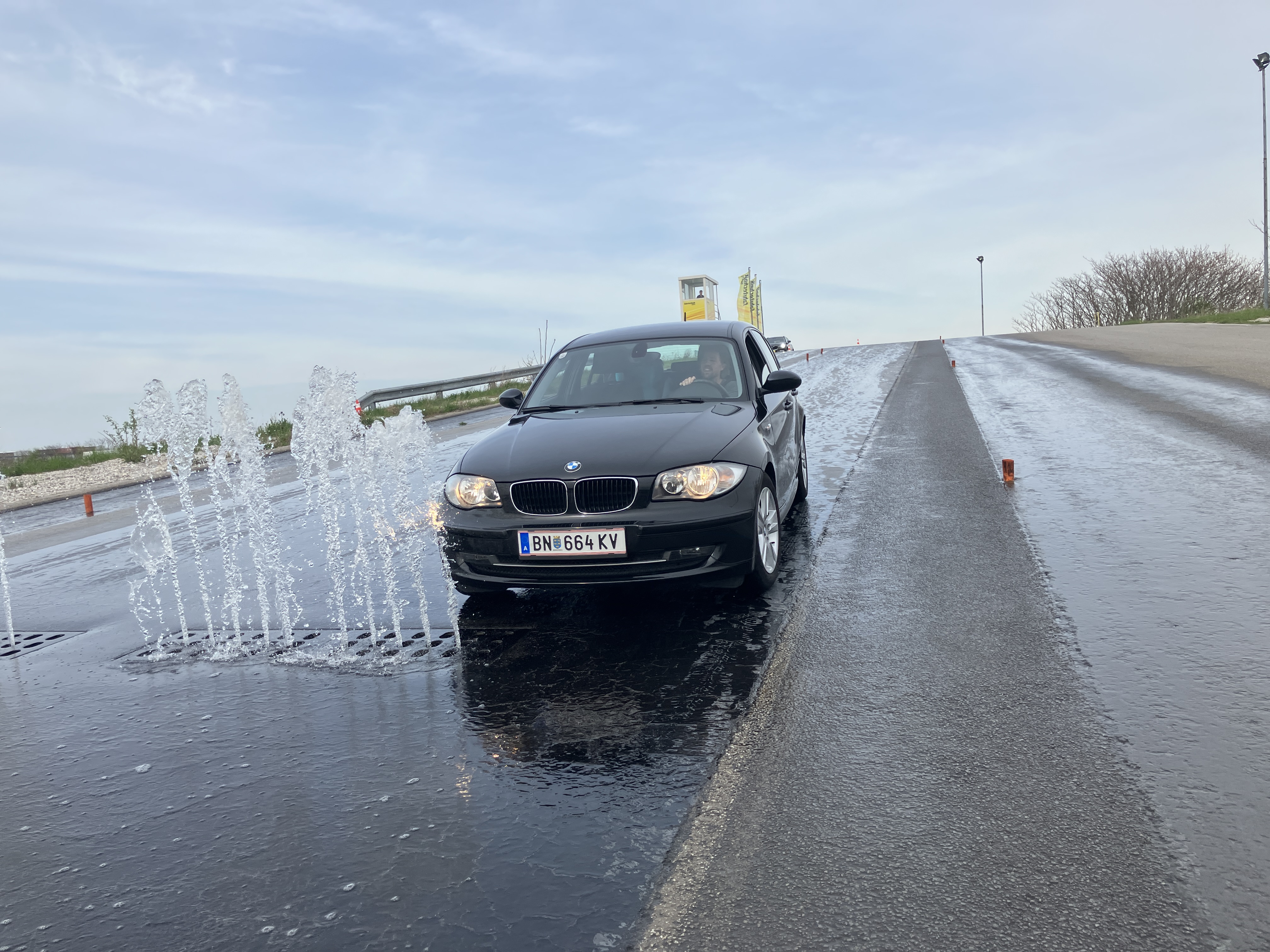 Auf dem Bild sieht man ein scwarzes Fahrzeug, das auf nasser Fahrbahn gerade ein Wasserhindernis umfährt.