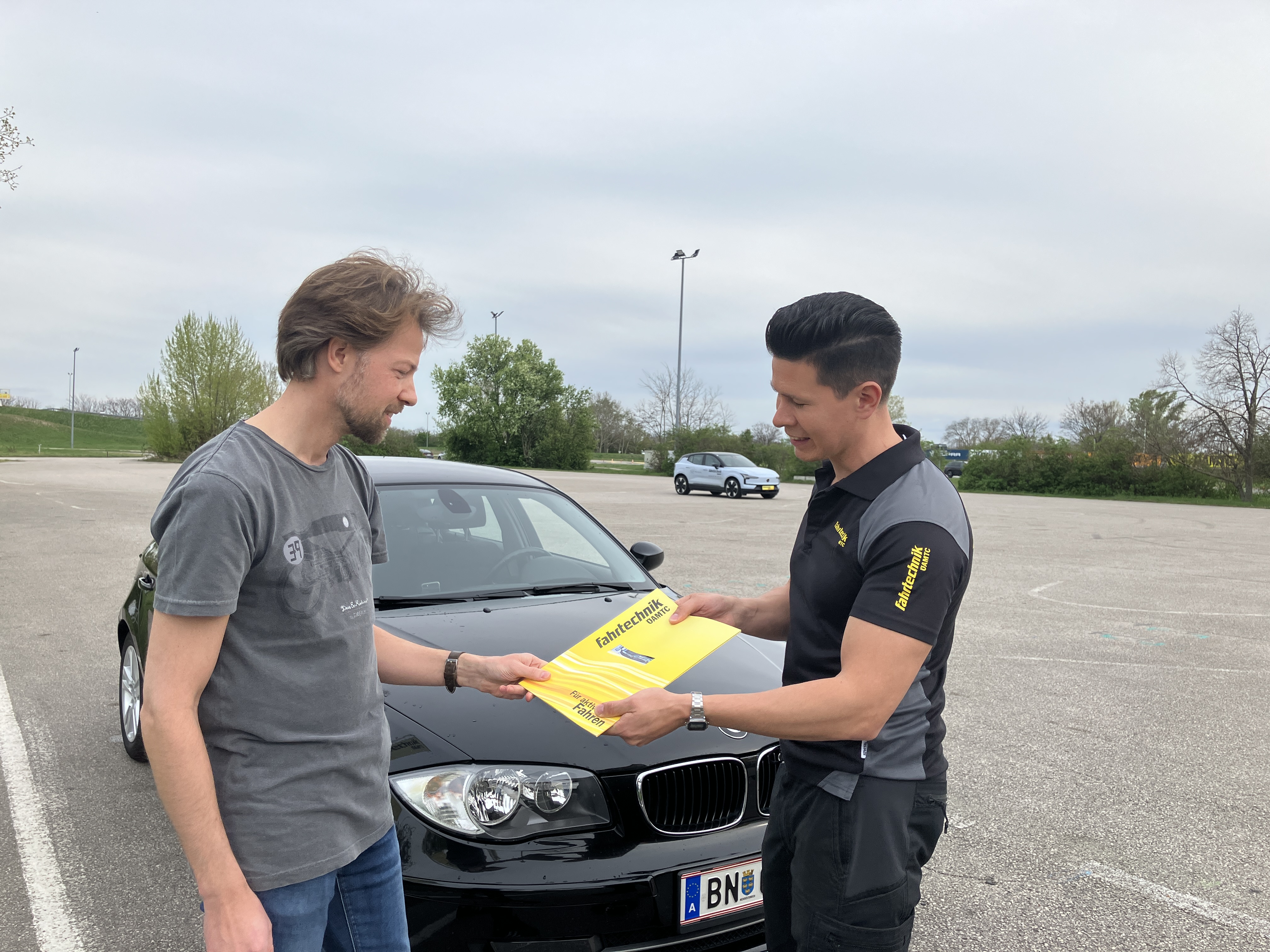 Auf dem Bild sieht man zwei Männer vor einem schwarzen Fahrzeug. Beide sind dunkel gekleidet. Der rechte überreicht dem linken eine gelbe Mappe.
